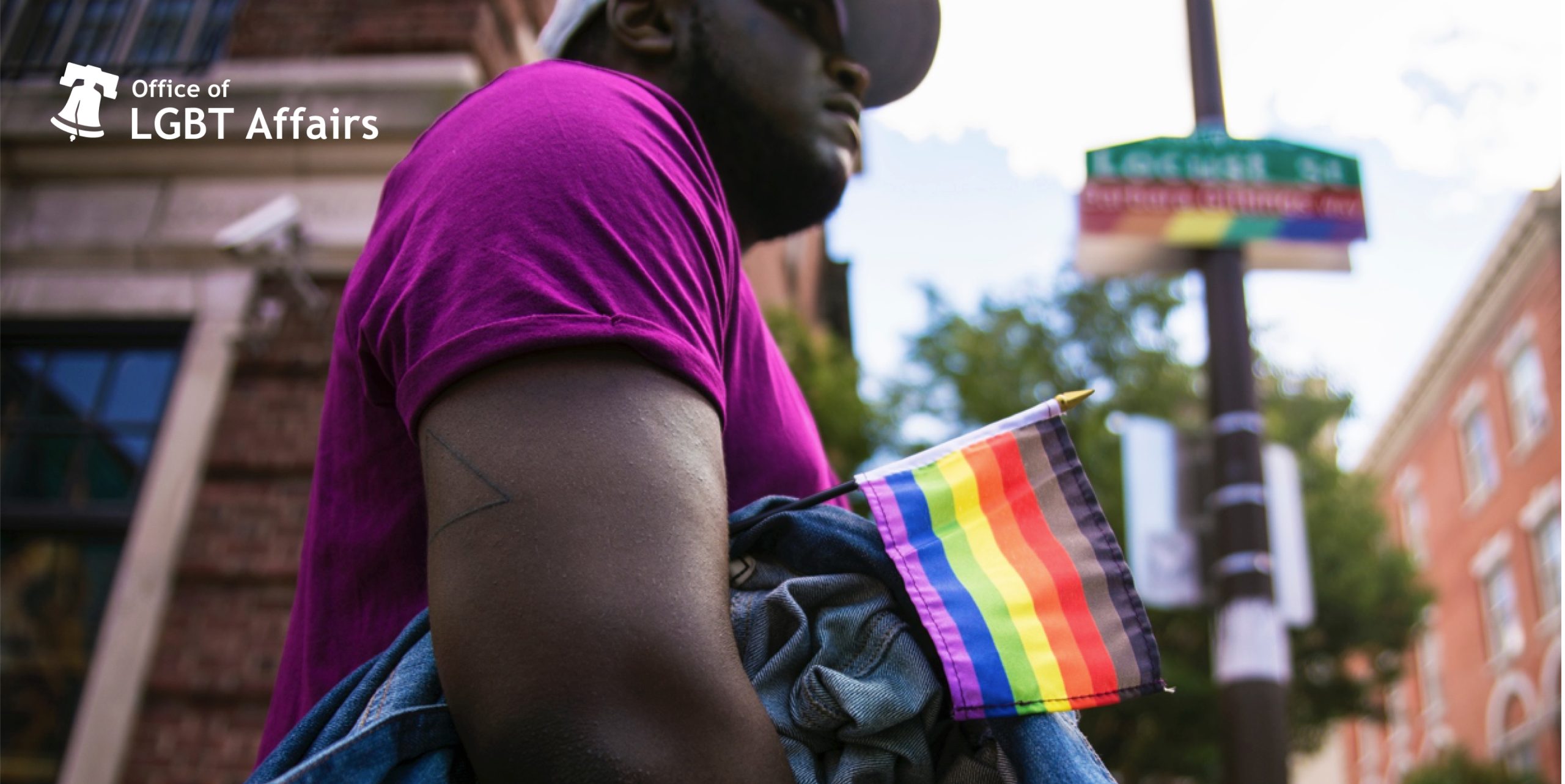 Close up of man on city street holding the new Tierney-created LGBTQ+ black, brown, red, orange, yellow, green, blue and purple flag under his arm